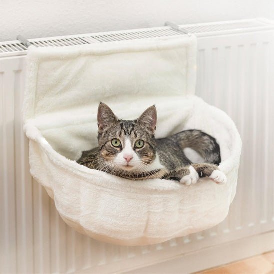 Chat blanc et gris détendu dans un hamac pour chat suspendu, fixé à un radiateur blanc