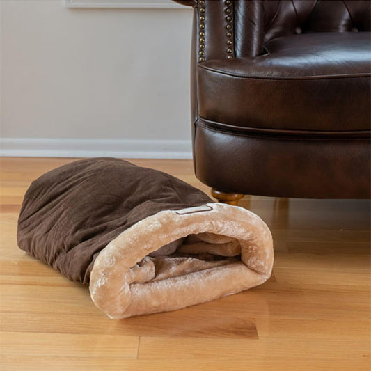 Sac de couchage pour chat marron et beige posé au sol près d'un fauteuil en cuir.