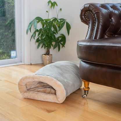 Sac de couchage pour chat gris et beige près d'un fauteuil en cuir et d'une plante.
