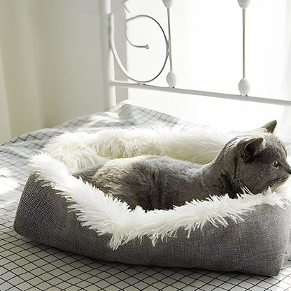 Coussin pour chat cocon Chadoux blanc, effet nid douillet posé sur un lit, avec un chat gris apaisé en lumière naturelle.