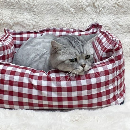 Lit pour chat rouge à carreaux avec un chat gris allongé, sur une couverture duveteuse.